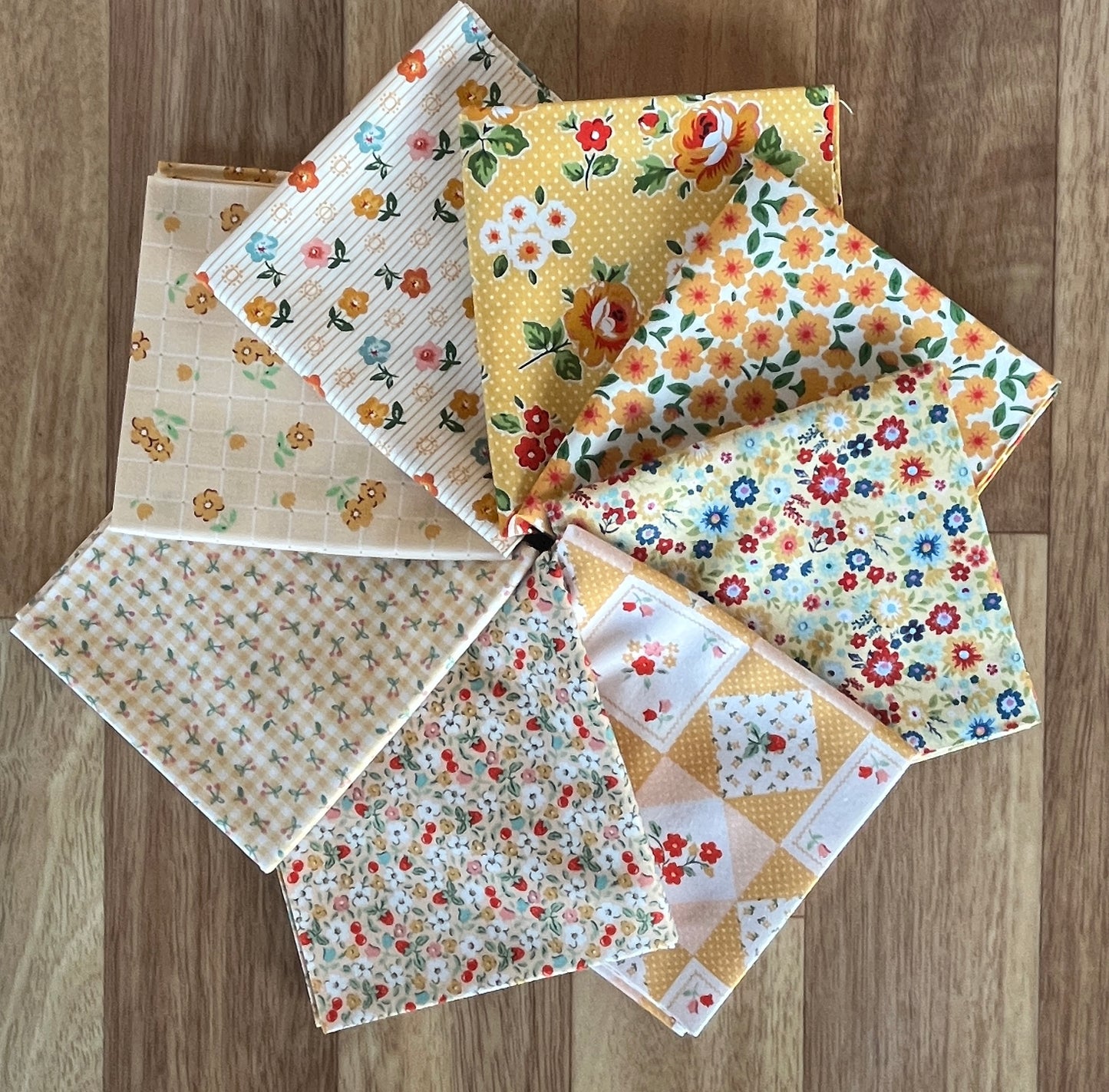 Six folded fabric squares with various floral and geometric patterns arranged in a fan shape on a wooden floor.