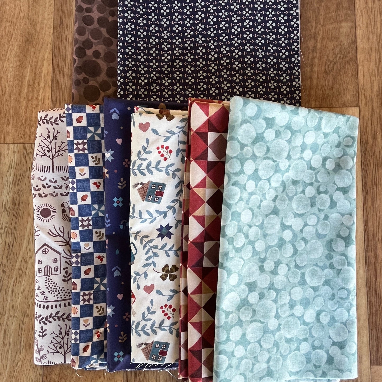 Five folded fabric pieces with various patterns and colors are arranged on a wooden floor. The fabrics include a brown animal print, a black and white floral pattern, a blue and white checkered pattern, a cream and brown floral pattern, and a light blue and white bubble pattern.